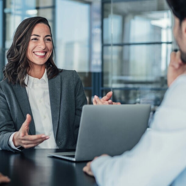 Eine lachende Frau im grauen Blazer sitzt vor einem Laptop an einem Tisch gegenüber zwei Personen in einem Raum mit Glaswänden.