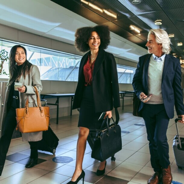 Drei Personen in eleganter Business-Kleidung mit Handgepäck laufen nebeneinander durch ein Flughafenterminal.