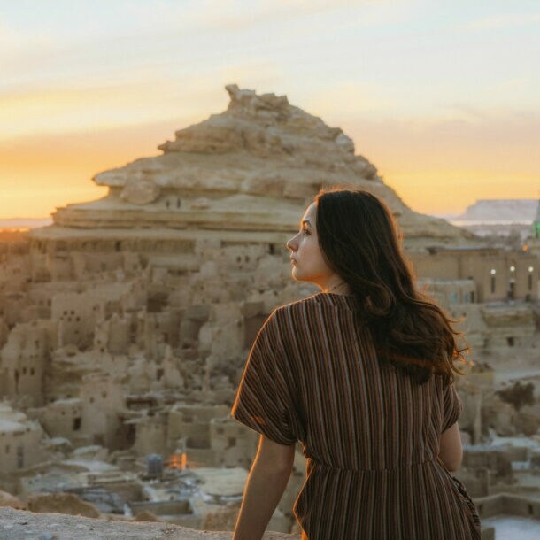 Frau mit braunem gestreiftem Kleid sitzt auf einer Mauer und blickt auf die Felsenstadt in der Siwa-Oase bei Sonnenuntergang in Ägypten.