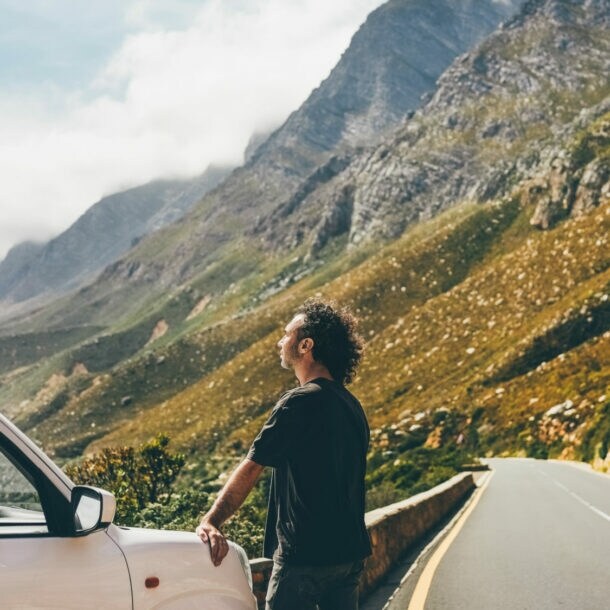 Mann lehnt an weißem Auto an einer Bergstraße, umgeben von steilen Felsen und bewölktem Himmel.