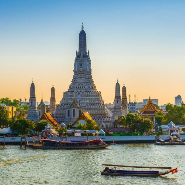 Vor einem großen Fluss mit Booten ragt ein großer Tempel empor, er ist umgeben von kleineren Türmen und Häusern mit spitzen Dächern.