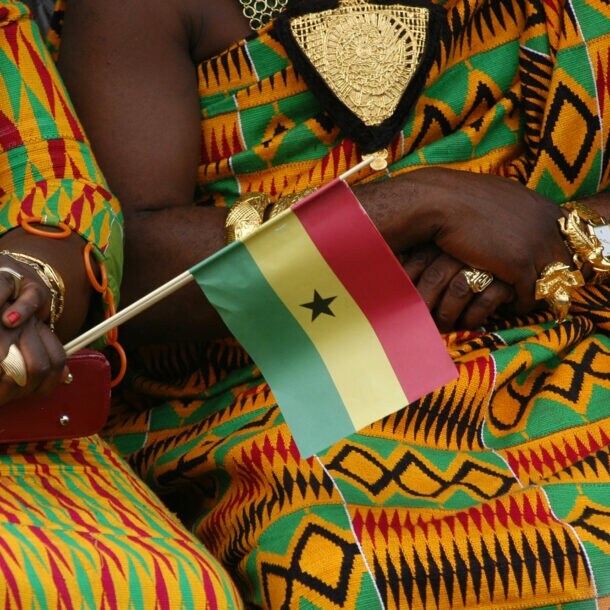 Oberkörper zweier sitzender Frauen in traditionellen ghanaischen Kleidern aus buntem Kente-Stoff und Goldschmuck, davor eine kleine ghanaische Flagge aus Papier in einer Hand gehalten.