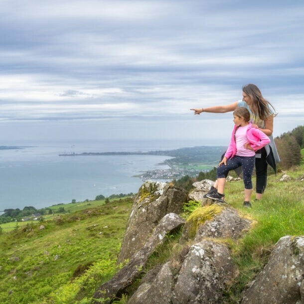 Eine Frau steht mit einem Mädchen auf einem Hügel in einer grünen Graslandschaft an einer Küste und zeigt hinaus aufs Meer.