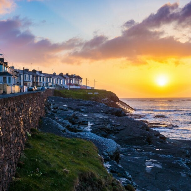 Sonnenuntergang über der Küste von Kilkee mit Reihenhäusern auf einer Steinmauer und Felsen am Meer