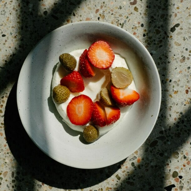Schale mit Joghurt, belegt mit geschnittenen Erdbeeren und kleinen grünen Früchten, auf marmoriertem Untergrund mit Schatten.