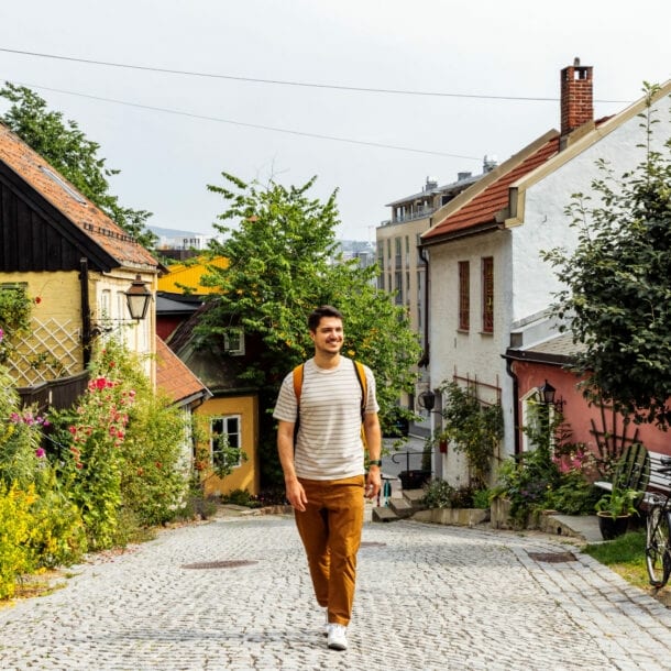 Person mit gestreiftem T-Shirt und brauner Hose geht auf gepflasterter Straße in Oslo, umgeben von bunten Häusern und Blumen.