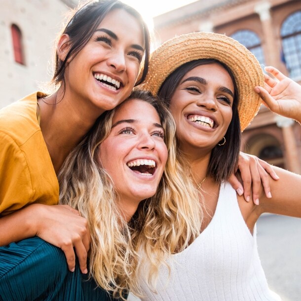 Drei lachende junge Frauen stehen sich umarmend auf einem Platz.