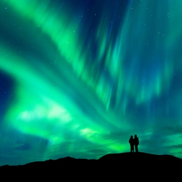 Zwei Personen stehen auf einem Hügel und beobachten das grün leuchtende Nordlicht am Nachthimmel in Norwegen.