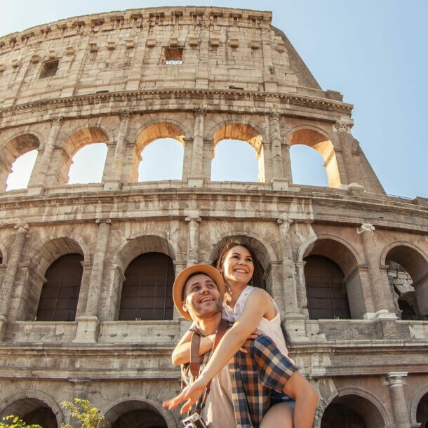 Das Kolosseum in Rom bei Sonnenlicht, davor ein Mann mit kariertem Hemd, der eine Person auf dem Rücken trägt.