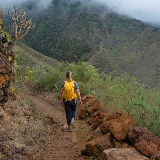 Eine Person mit gelbem Rucksack geht auf einem Pfad in bergiger Landschaft auf Teneriffa. Der Himmer ist bewölkt, am Wegesrand ist grüne Vegetation.