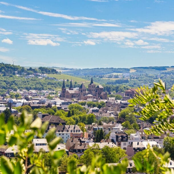 Blick auf Trier mit der Porta Nigra und umliegenden Gebäuden, im Vordergrund grüne Blätter, im Hintergrund bewaldete Hügel unter blauem Himmel mit Wolken.