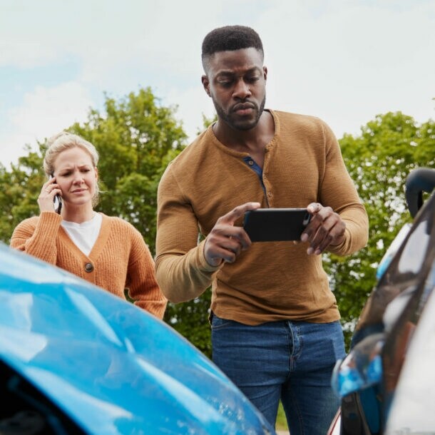 Ein Mann und einer Frau mit Smartphones stehen hinter zwei Autos mit Blechschäden, er macht ein Foto vom Schaden.