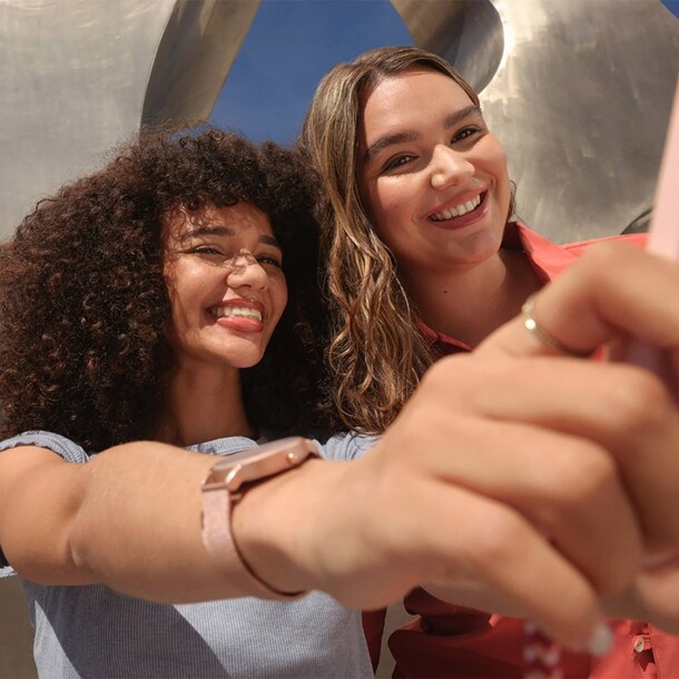 Zwei junge Frauen machen ein Selfie mir einem rosafarbenen Smartphone vor einer Metallskulptur unter freiem Himmel.