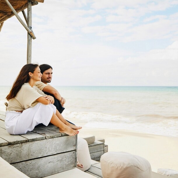 Ein Paar sitzt entspannt auf einer Holztribüne mit Polstermöbeln an einem weißen Sandstrand und blickt aufs Meer.