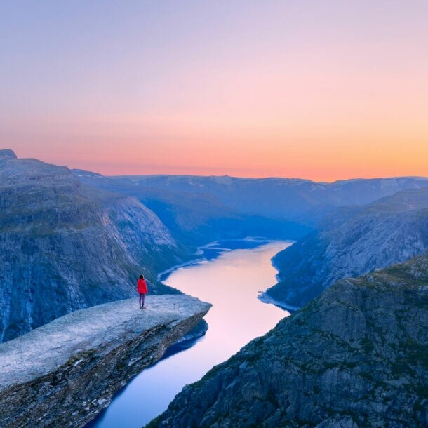 Bergklippe Trolltunga mit Person in roter Jacke, die auf dem Felsvorsprung über einem tiefen Fjord steht, umgeben von Bergen im Abendlicht.