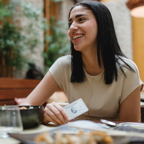 Eine Frau sitzt neben einer anderen Person an einem gedeckten Tisch und hält eine Platinum Plus Card von American Express in der Hand.