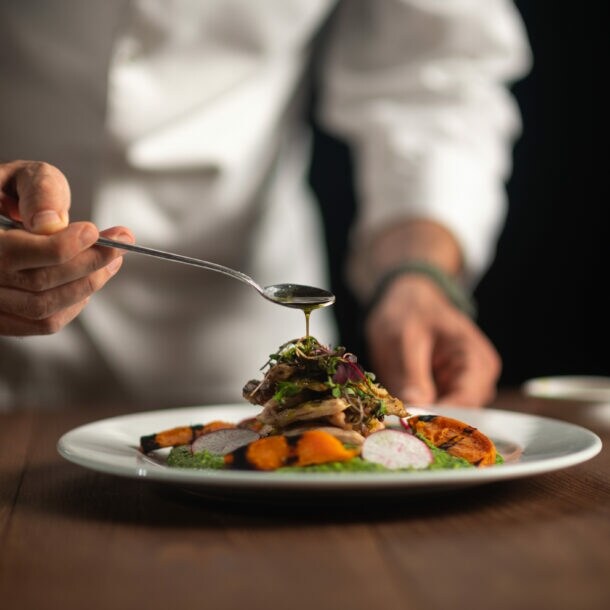 Die Hände eines Kochs, der Soße aus dem Löffel auf eine Mahlzeit auf einem weißen Teller gießt.