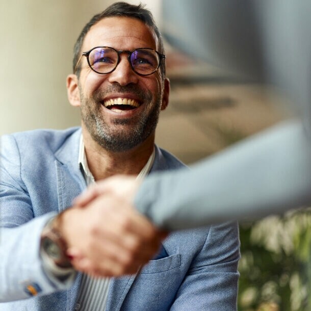 Ein lächelnder Mann im hellblauen Sakko schüttelt einer Person die Hand.