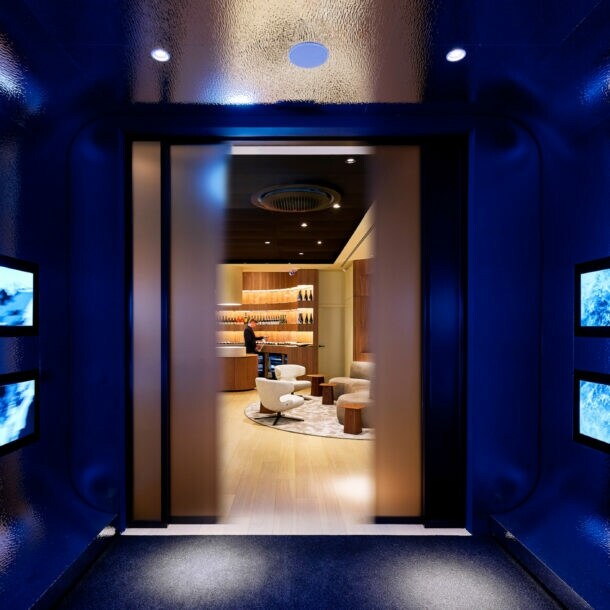 Blick durch einen dunklen, blau beleuchteten Gang mit vier rechteckigen Bildschirmen an den Seiten auf einen hellen, modernen Raum mit Sesseln und Regalen.