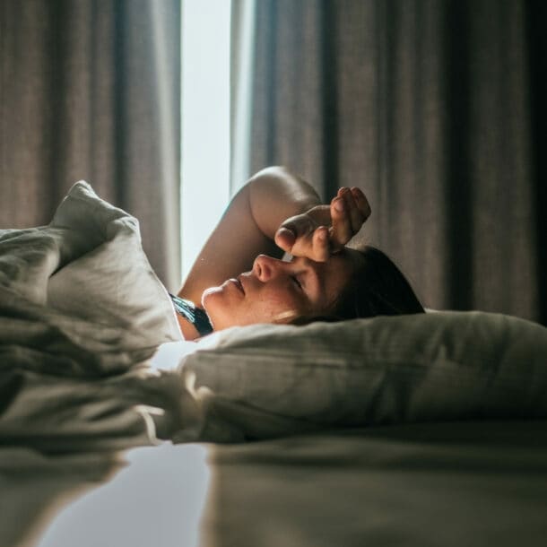 Eine Person liegt auf einem Bett, mit einem weißen Bettlaken und Kissen. Licht strömt durch ein Fenster, das von Vorhängen umgeben ist.