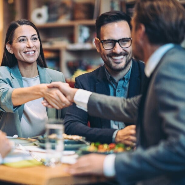 Eine lächelnde, elegant gekleidete Frau im Blazer und ein Mann im Anzug geben sich die Hand an einem Tisch mit zwei weiteren Personen.