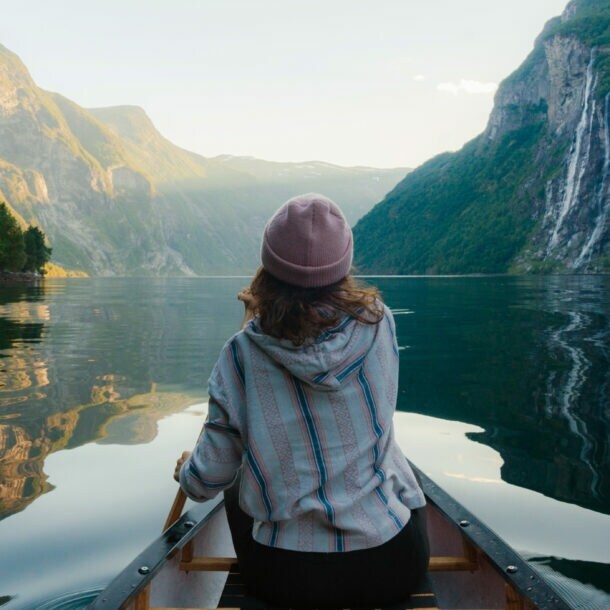 Person mit rosa Mütze und gestreiftem Pullover sitzt in einem Kanu auf ruhigem Wasser, umgeben von hohen, bewaldeten norwegischen Fjordwänden mit Wasserfall.