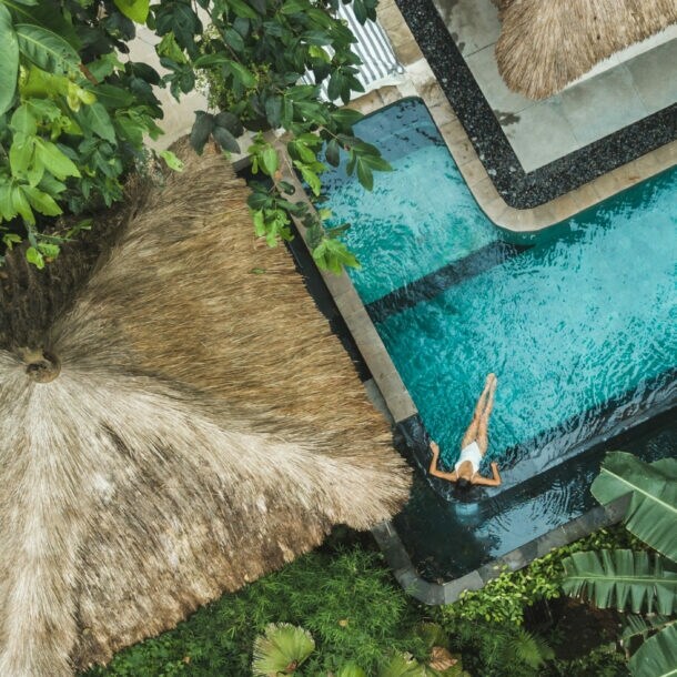 Eine Person schwimmt in einem blauen Pool, umgeben von tropischen Pflanzen und einem Strohdach. Der Blick ist von oben.