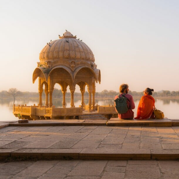 Zwei Personen mit Rucksäcken sitzen in einer buddhistischen Tempelanlage an einem See im weichen Sonnenlicht.