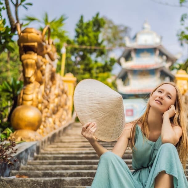 Eine Frau sitzt lächelnd auf einer Treppe. Sie schwenkt einen traditionellen vietnamesischen Hut. Im Hintergrund sind ein Tempel und goldene Statuen zu sehen.
