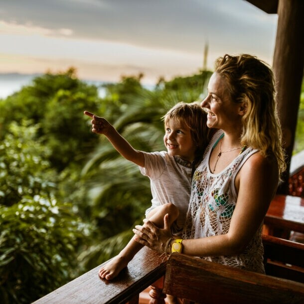 Eine Frau hat einen kleinen Jungen im Arm, beide blicken von einer Holzterrasse in den Dschungel von Thailand, hinter ihnen das Meer.
