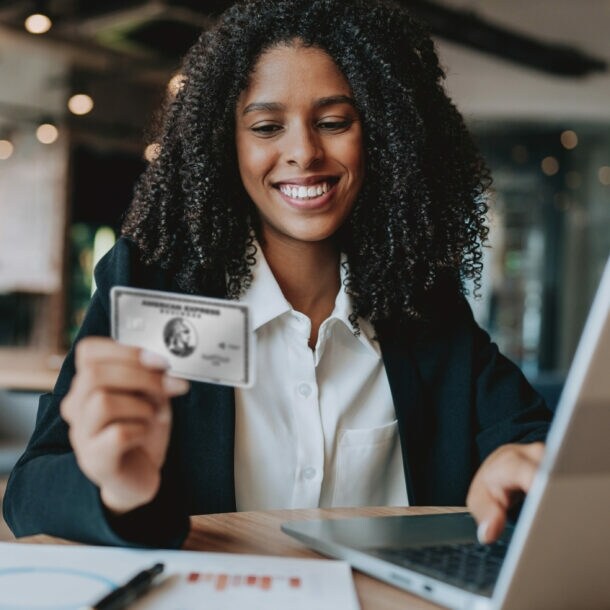 Eine lächelnde Frau bedient mit einer Hand einen Laptop und hält in der anderen Hand eine silberne Kreditkarte von American Express.