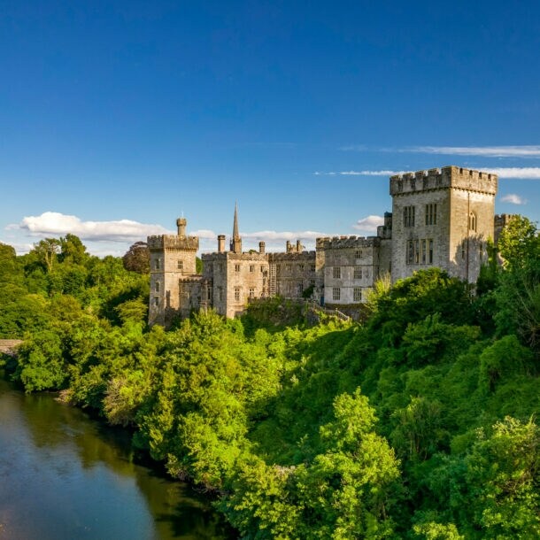 Luftaufnahme von Lismore Castle in Waterford, umgeben von dichtem Grün und einem Fluss mit einer Steinbrücke im Vordergrund.