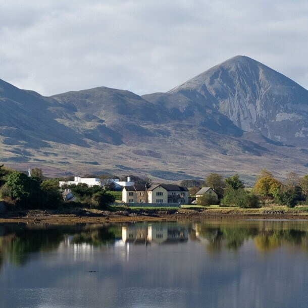 Vor einem großen Gewässer stehen kleine Häuser und Bäume, in Hintergrund ragt ein großer Berg empor, die Szene spiegelt sich in der Wasseroberfläche.