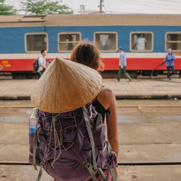 Eine Person mit einem großen, runden Strohhut und einem lila Rucksack steht auf einem Bahnsteig und schaut auf einen Zug. Im Hintergrund sind mehrere Personen und ein Zug zu sehen.