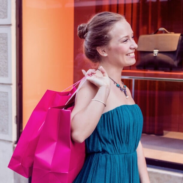 Eine lächelnde Frau im Kleid steht vor dem Schaufenster einer Modeboutique und hält zwei pinke Einkaufstüten über ihrer Schulter.