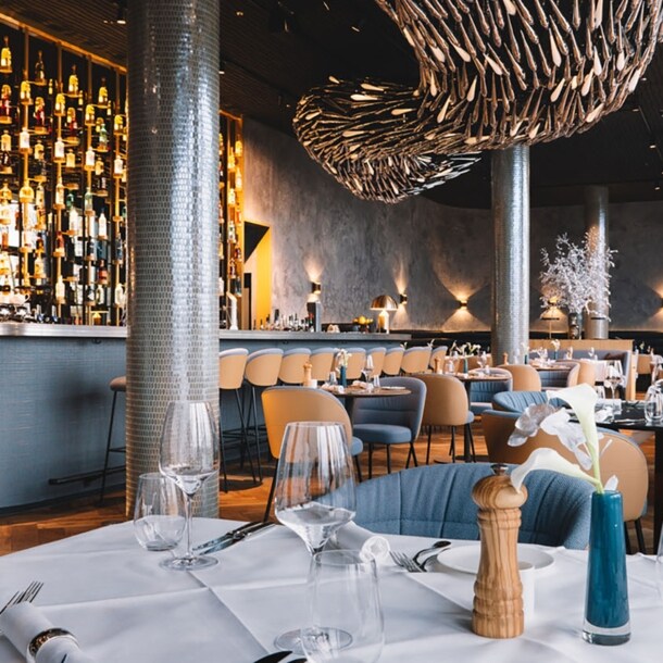 Stilvoller, modern eingerichteter Speisesaal mit Barbereich und eingedeckten Tischen unter einer abstrakten Deckenskulptur.