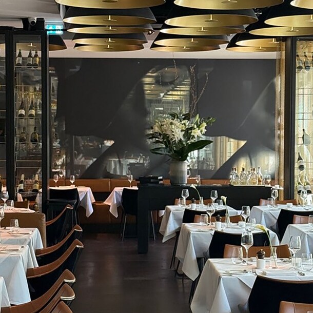 Eleganter Speiseraum mit weiß eingedeckten Tischen und goldfarbenen Deckenlampen vor einem schwarzen Wandbild.