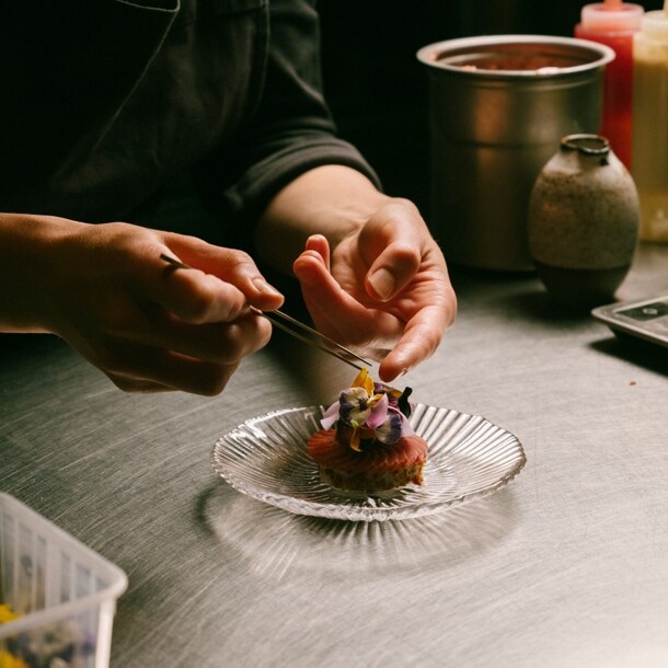 Zwei Hände garnieren eine edle Vorspeise mit Blüten auf einer Küchentheke.