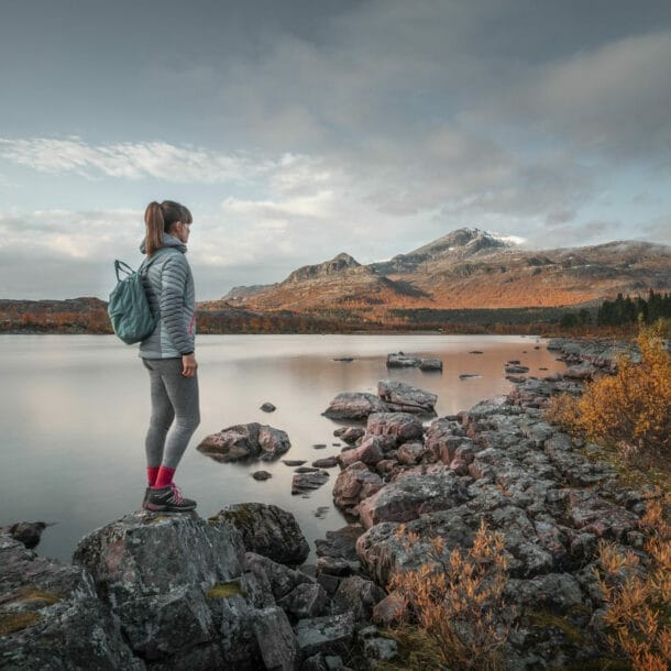 Eine Frau mit Rucksack steht auf einem Stein an einem See in flacher Felslandschaft mit herbstlicher Vegetation.