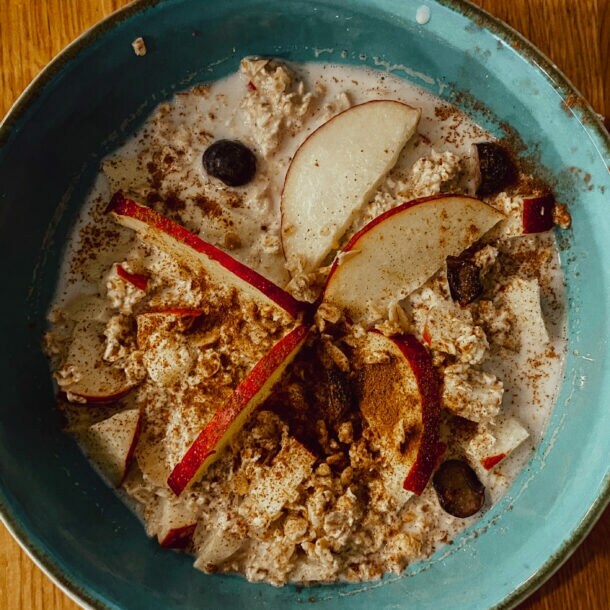 Blaue Schüssel mit Porridge, garniert mit Apfelscheiben, Blaubeeren und Zimt, auf Holztisch mit Glas Wasser und Serviette.