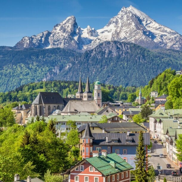 Blick auf eine Stadt mit historischen Gebäuden und einer Kirche vor bewaldeten Hügeln und schneebedeckten Bergen im Hintergrund.