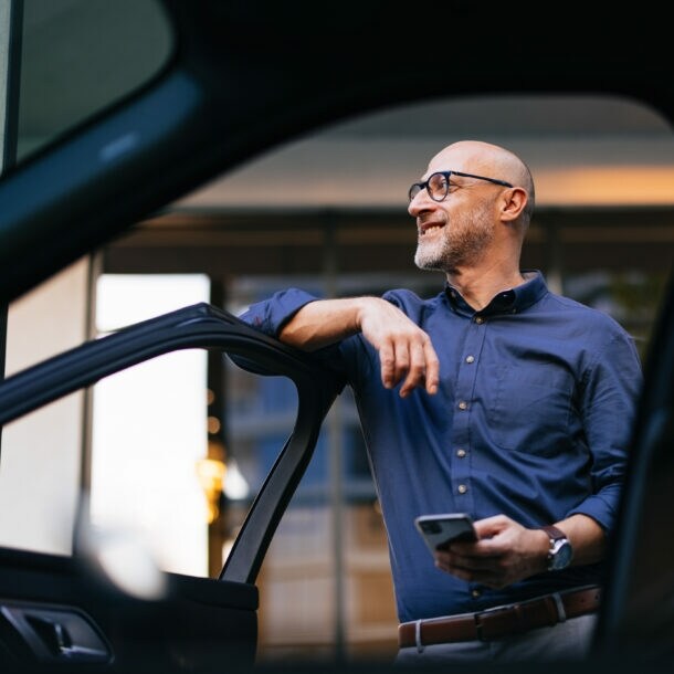 Ein Mann im blauen Hemd und mit einem Smartphone in der Hand lehnt an einer geöffneten Autotür und blickt lächelnd in die Ferne.