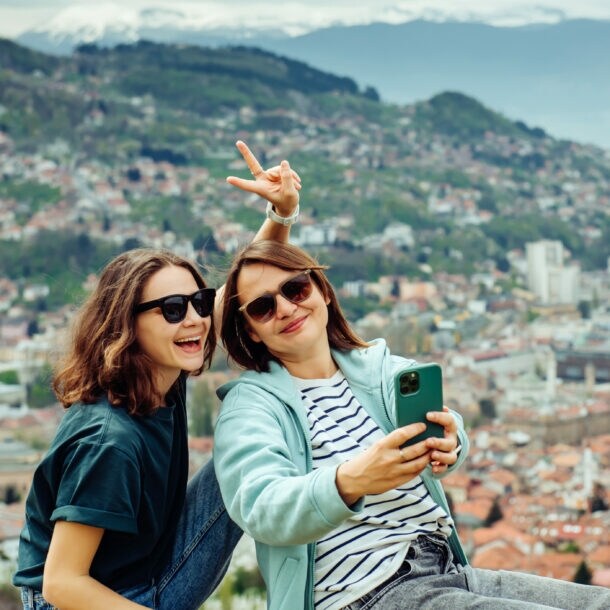 Zwei junge Frauen sitzen auf einer Mauer mit Blick auf eine Stadt, eine macht ein Selfie mit einem Smartphone und zeigt ein Peace-Zeichen.