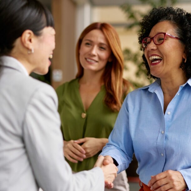 Vier Personen stehen beieinander in einem Büro, im Vordergrund geben sich zwei lachende Frau die Hand.