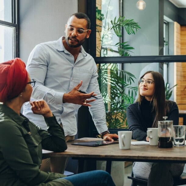 Mehrere Personen sitzen oder stehen an einem Tisch und schauen eine Person an, die spricht.