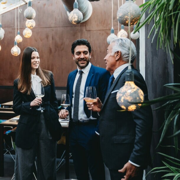 Personen in eleganter Kleidung stehen mit Weingläsern in den Händen beieinander in einem elegant geschmückten Raum.