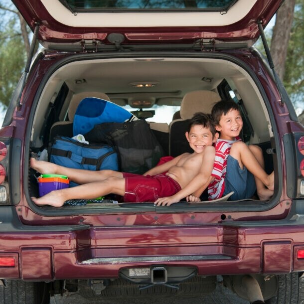 Zwei Jungen in Strandoutfits sitzen fröhlich in einem offenen Kofferraum, hinter ihnen das Meer.