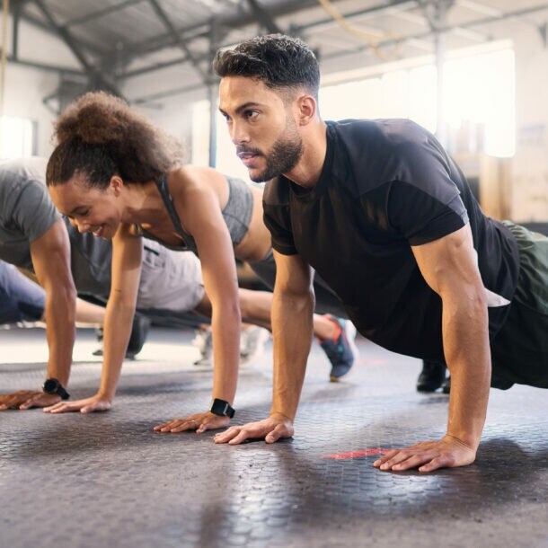 Mehrere Personen in Sportkleidung machen Liegestütze in einem hellen Fitnessstudio mit Gummiboden.