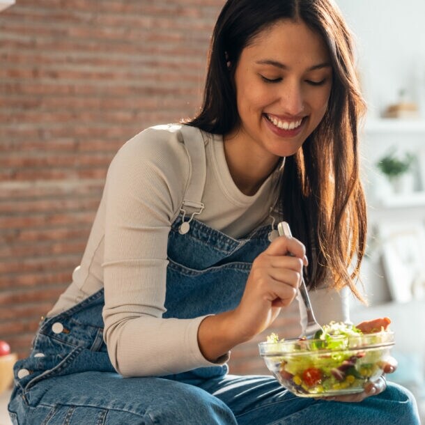 Frau in Jeanslatzhose sitzt auf einem Tisch und isst einen Salat aus einer Glasschüssel mit einer Gabel.