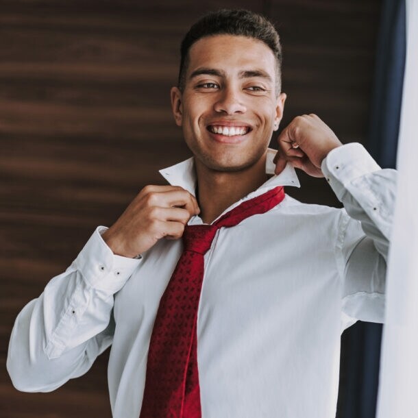 Ein junger Mann im weißen Hemd mit roter Krawatte richtet sich lächelnd den Kragen vor einem Spiegel.
